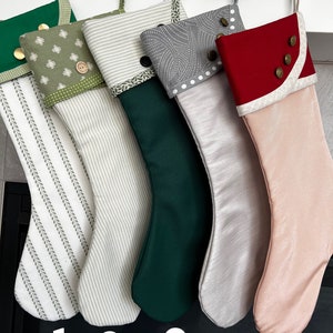 May include: Five Christmas stockings in various colors and patterns hang from wooden pegs. The stockings include green, white, and red fabrics with decorative buttons and patterned cuffs. Numbers 1 through 5 are printed at the bottom.