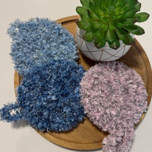 May include: Two fluffy, textured washcloths in blue and pink with silver threads, displayed on a wooden tray. A small succulent plant in a white and gold geometric patterned pot sits nearby. The washcloths have a loop for hanging.