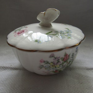 Op de afbeelding: Een witte porseleinen sieradenschaal met deksel, versierd met een bloemenpatroon in roze, blauw en geel. Het deksel heeft een vlindervormige handgreep en gouden rand. De schaal heeft een geschulpte rand.