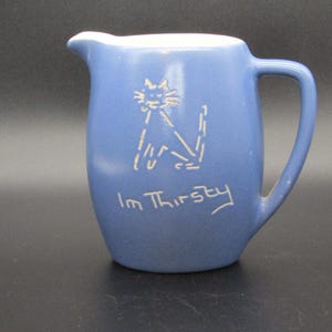 May include: A blue ceramic pitcher with a handle and spout. A white cat illustration and the words "I'm Thirsty" are on the front. The pitcher is likely used for serving beverages.