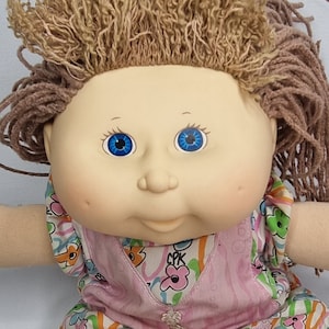Può includere: Una bambola Cabbage Patch Kid con capelli biondi, occhi azzurri e un vestito floreale rosa e bianco.