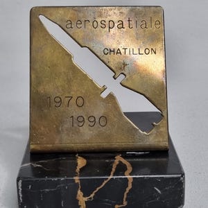 May include: A vintage brass desk ornament with the text "aerospatiale CHATILLON" and the years "1970 1990". The brass piece has a cut-out design of a rocket or missile shape, mounted on a black and gold marble base.