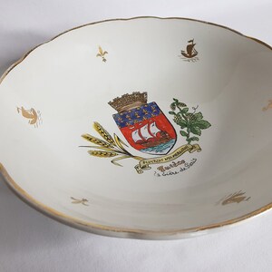 May include: White ceramic bowl with a gold rim and a coat of arms design in the center. The coat of arms features a red shield with a white ship, a blue and gold crown, and a blue and gold banner with the text "Fluctuat Nec Mergitur". The bowl also features a gold fleur-de-lis design and the text "Foulées à bière de Sois".