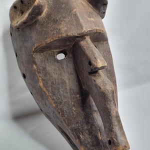 Puede incluir: Una máscara de madera marrón oscuro con una cara humanoide y dos pequeños cuernos en la parte superior. La máscara tiene una boca grande abierta y un pequeño agujero para un ojo.