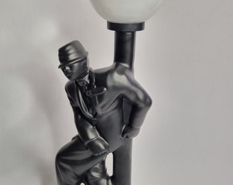 May include: Black ceramic table lamp featuring a stylized figure of a man leaning against a lamp post. The lamp has a white globe shade.