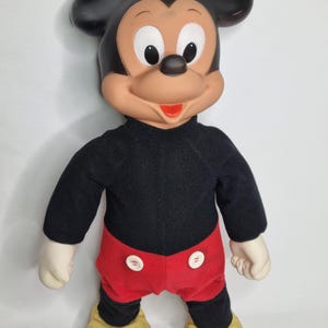 Könnte beinhalten: Ein Vintage-Plüschtier Mickey Mouse mit schwarzen Ohren, einem schwarzen und roten Outfit und gelben Schuhen. Das Spielzeug hat ein lächelndes Gesicht mit schwarzen Augen und einem roten Mund.