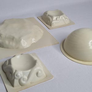May include: Four white plastic molds for making decorative concrete items. The molds are shaped like a rock, a dome, and two different types of bases.