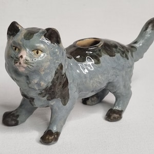 May include: A ceramic cat figurine with a glossy finish, featuring a light blue-gray base color with dark brown patches on the head, back, and paws. The cat has a pink nose, yellow eyes, and a circular opening on its back, with a raised tail.