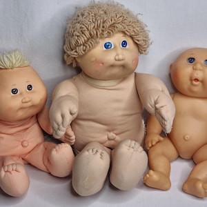 Può includere: Tre bambole vintage Cabbage Patch Kids. Le bambole hanno corpi in tessuto morbido e teste in vinile con tratti del viso dipinti. Le bambole hanno acconciature e tonalità della pelle diverse. Le bambole sono sedute su una superficie bianca.