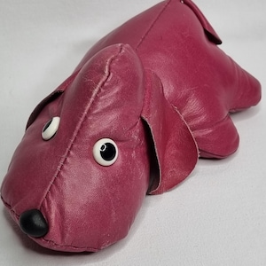 May include: A plush, burgundy-coloured dog toy with black button eyes and a black nose. The toy is made of soft, textured leather.