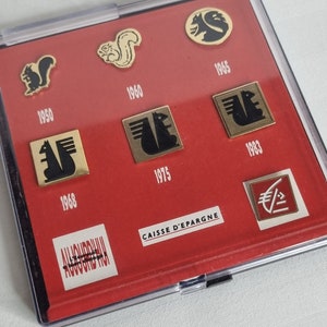 May include: A red display case containing a collection of vintage pins featuring a squirrel logo. The pins are arranged in chronological order from 1950 to 1983. The case also includes a label that reads "CAISSE D'EPARGNE".