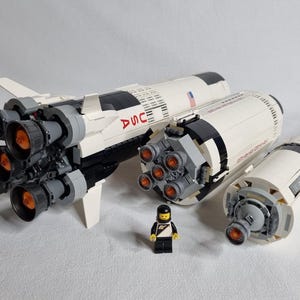 May include: A white and black Lego model of the Saturn V rocket with a small astronaut minifigure. The rocket has multiple stages and detailed engines.