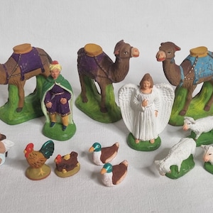 May include: A collection of miniature figurines depicting a nativity scene. The set includes camels, an angel, a king, sheep, ducks, a rooster, and a hen. The figurines are painted in various colors, including brown, green, white, and purple.