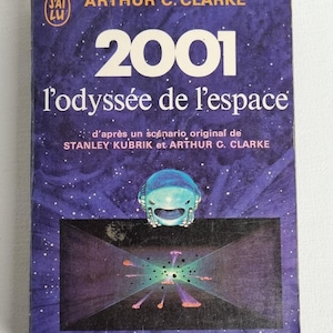 Peut inclure: Une couverture de livre violette avec le titre "2001 l'odyssée de l'espace" par Arthur C. Clarke. La couverture présente une illustration bleue et blanche d'un vaisseau spatial et d'un champ d'étoiles.