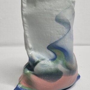 May include: A handmade ceramic vase with a unique, abstract design. The vase features a light blue and white upper section with flowing blue and green accents. The base is a blend of pink, green, and blue, creating a colorful, artistic piece.