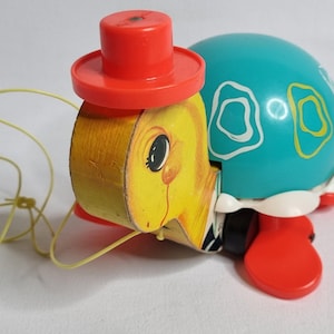 May include: A vintage pull-along toy turtle with a yellow head, a teal shell, and red feet. The turtle wears a red top hat and has a yellow pull string. The shell has white and yellow decorative shapes.