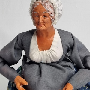 May include: A handcrafted figurine of an elderly woman, dressed in a gray dress with a white collar and a white lace bonnet. The doll has painted features and orange-toned hands. The skirt has a blue floral pattern.