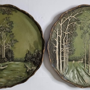Puede incluir: Dos platos decorativos redondos con un diseño de paisaje en relieve en tonos verde oliva y marrón oscuro. El plato izquierdo muestra una escena fluvial con árboles, mientras que el derecho presenta un camino arbolado hacia una pequeña casa.
