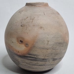 Może przedstawiać: Sferyczny wazon ceramiczny o matowej, jasnobeżowej powierzchni z subtelnymi szarymi poziomymi prążkami i wyraźną okrągłą pomarańczowo-brązową plamą. Naczynie posiada mały, wąski okrągły otwór w górnej części oraz gładki, zaokrąglony korpus.
