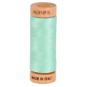 May include: A spool of mint green thread with wooden ends. The top wooden end has the word "AURIFIL" printed on it. The bottom wooden end has "MADE IN ITALY" printed on it. The thread is tightly wound around the spool.