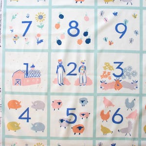 May include: A white fabric with a grid pattern featuring numbers 1-9 in blue. Each square has a different illustration, including a barn, farmers, sheep, and chickens. The fabric has a light blue border around each square.