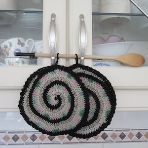 Op de afbeelding: Twee zwarte en witte gehaakte ovenwanten met een spiraalvormig ontwerp in roze, groen en beige, hangend aan een houten lepelsteun.