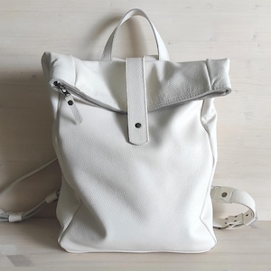 May include: White leather backpack with a roll-top design. Features a top handle, adjustable shoulder straps, and a front flap secured with a snap closure. The bag has a zipper closure and a textured surface.