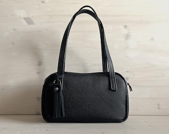 May include: A black handbag with a textured surface, featuring two long shoulder straps and a decorative tassel attached to a silver-toned metal ring on the front. The bag has a rectangular shape with rounded corners and a zipper closure.