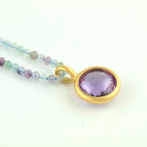 Puede incluir: Un collar de oro con un colgante de amatista púrpura. El collar está hecho de pequeñas cuentas redondas en tonos de azul, verde y morado.