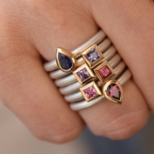 Könnte beinhalten: Eine Sammlung von Silberringen mit goldenen Akzenten, jeweils mit einem anderen Edelstein besetzt. Die Edelsteine sind in verschiedenen Farben gehalten, darunter Blau, Lila und Rosa. Die Ringe sind auf einem Finger gestapelt und zeigen die Vielfalt der Designs und Farben.