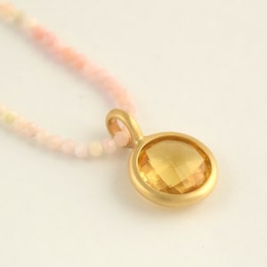 Puede incluir: Un collar de oro con un colgante redondo de piedra preciosa amarilla. El collar está hecho de pequeñas cuentas rosas.
