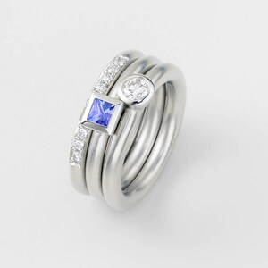 Puede incluir: Un conjunto de tres anillos de plata con una piedra preciosa cuadrada azul y una piedra preciosa redonda blanca. Los anillos est&aacute;n apilados uno encima del otro.