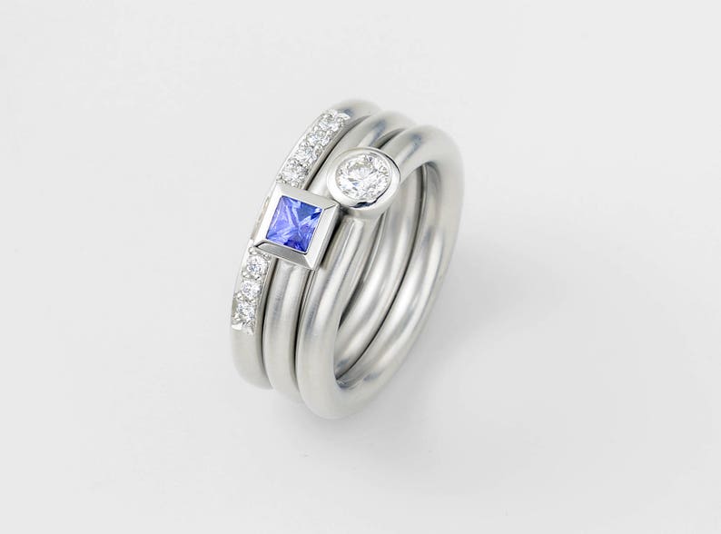 Puede incluir: Un conjunto de tres anillos de plata con una piedra preciosa cuadrada azul y una piedra preciosa redonda blanca. Los anillos est&aacute;n apilados uno encima del otro.