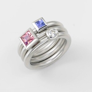 Puede incluir: Un anillo de plata con tres piedras preciosas: un cuadrado rosa, un cuadrado azul y una piedra redonda transparente.