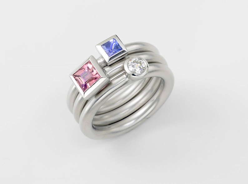Puede incluir: Un anillo de plata con tres piedras preciosas: un cuadrado rosa, un cuadrado azul y una piedra redonda transparente.