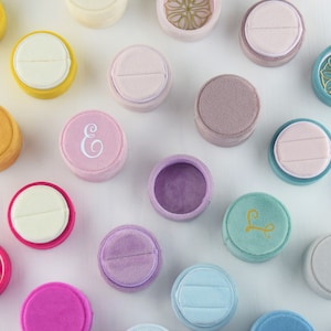 Puede incluir: Surtido de cajas de anillos redondas de terciopelo en varios colores, incluyendo rosa, azul, morado, amarillo y marrón. Algunas cajas tienen un diseño simple, mientras que otras presentan una letra grabada o un patrón decorativo.