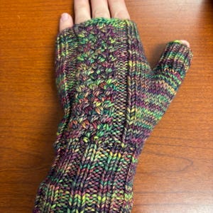 以下が含まれることがあります： 手編みの指なし手袋。手袋は、紫、緑、黄色、赤の混色で模様編みされています。親指は覆われていますが、指は露出しています。編み物の質感がよくわかります。