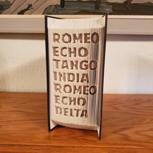 Puede incluir: Un libro blanco con las palabras "ROMEO ECHO TANGO INDIA ROMEO ECHO DELTA" dobladas en las p&aacute;ginas. El libro est&aacute; de pie sobre una superficie de madera.