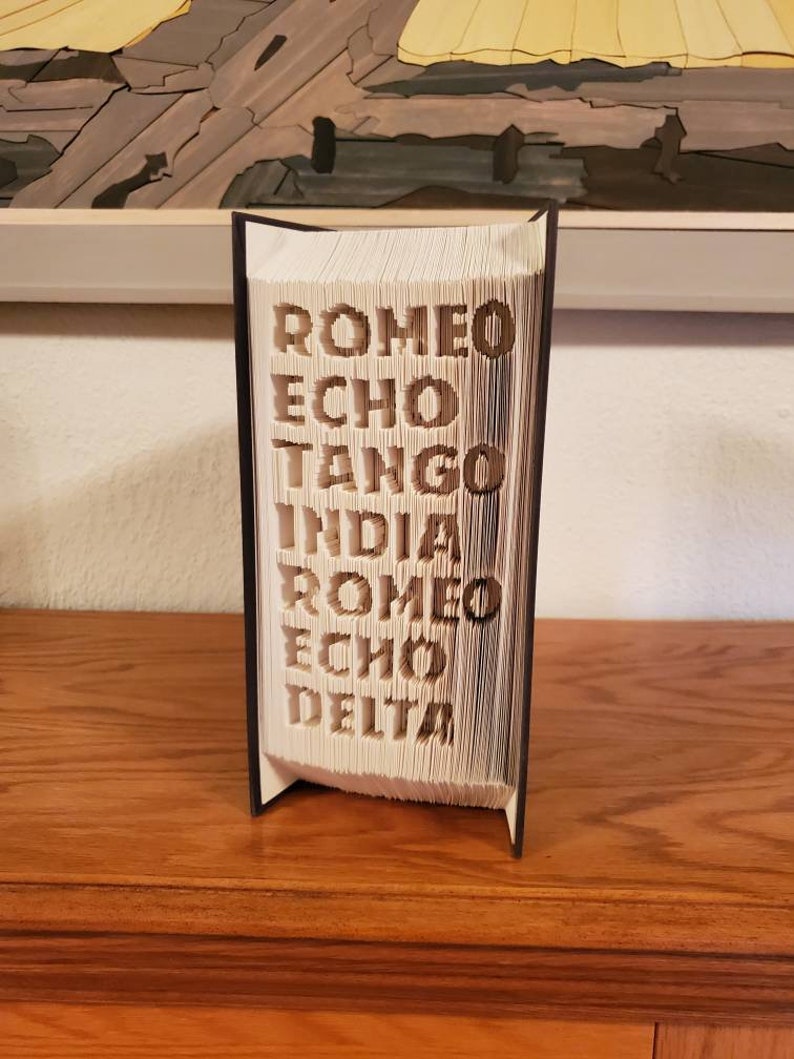 Puede incluir: Un libro blanco con las palabras "ROMEO ECHO TANGO INDIA ROMEO ECHO DELTA" dobladas en las p&aacute;ginas. El libro est&aacute; de pie sobre una superficie de madera.