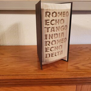 Puede incluir: Un libro con las palabras "ROMEO ECHO TANGO INDIA ROMEO ECHO DELTA" dobladas en las p&aacute;ginas. El libro est&aacute; de pie sobre una superficie de madera.
