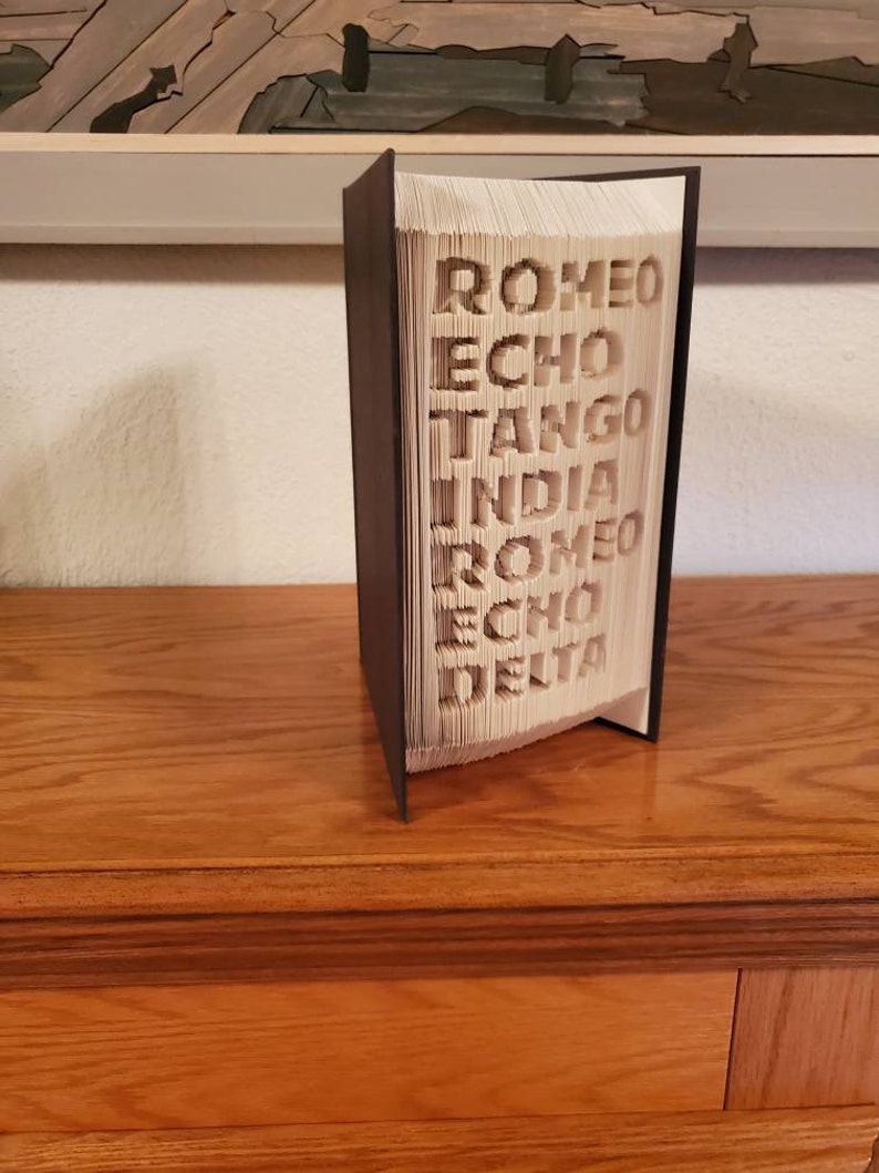 Puede incluir: Un libro con las palabras "ROMEO ECHO TANGO INDIA ROMEO ECHO DELTA" dobladas en las p&aacute;ginas. El libro est&aacute; de pie sobre una superficie de madera.