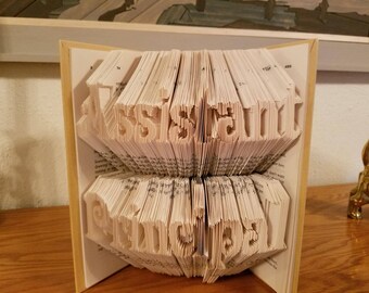 May include: A handmade book sculpture with the word "JUST MARRIED" cut into the pages. The book is open, revealing the carved letters in a stacked arrangement. The pages are beige, and the edges are visible.