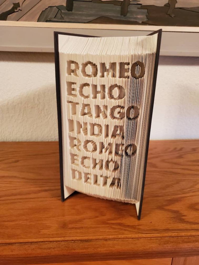 Puede incluir: Un libro con las palabras "ROMEO ECHO TANGO INDIA ROMEO ECHO DELTA" dobladas en las p&aacute;ginas. El libro est&aacute; de pie sobre una superficie de madera.