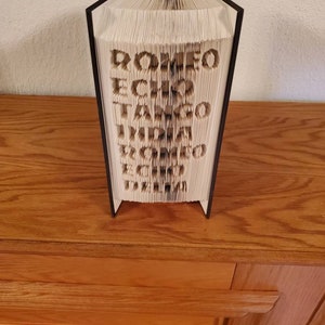 Puede incluir: Un libro con las palabras "ROMEO ECHO TANGO INDIA DELTA ZULU ECHO" doblado en una escultura 3D.