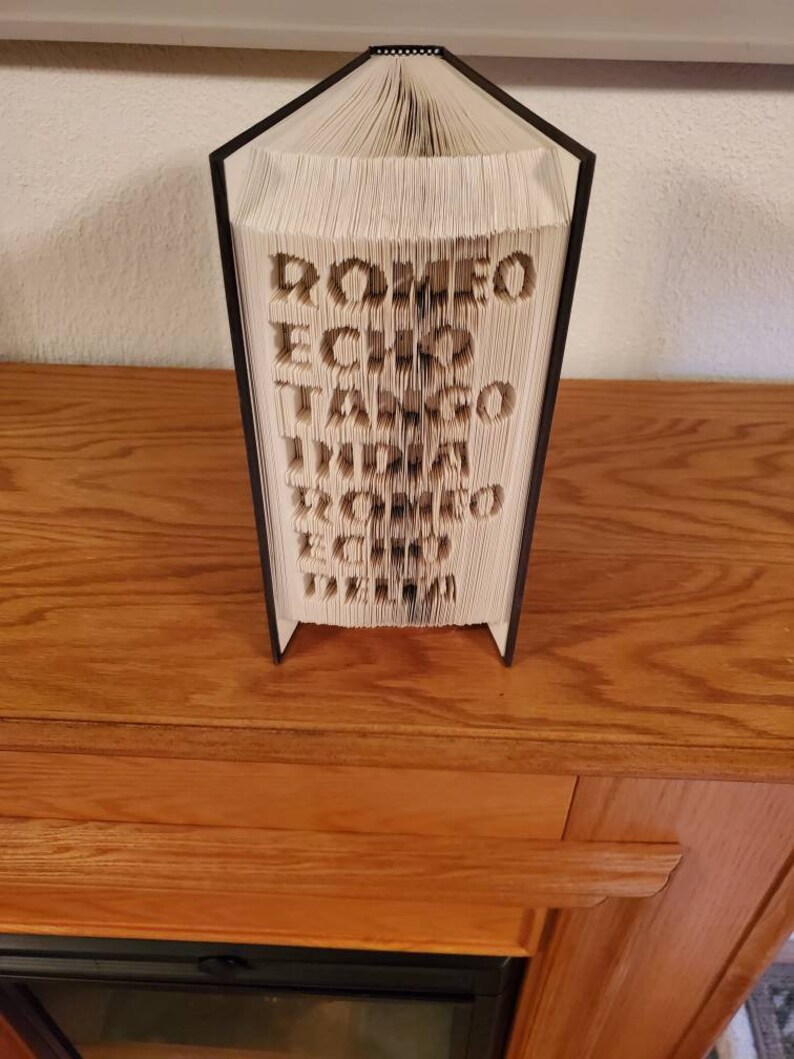 Puede incluir: Un libro con las palabras "ROMEO ECHO TANGO INDIA DELTA ZULU ECHO" doblado en una escultura 3D.