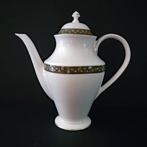 May include: A white porcelain teapot with a lid and a curved handle. The teapot has a decorative band of brown and green leaves around the middle and on the lid. The spout is long and slender. The teapot sits on a black surface.