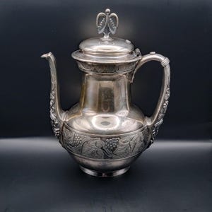 May include: Silver-toned antique coffee pot with a lid and handle. The pot features ornate grape and leaf detailing around the base and handle. The lid is topped with a decorative finial. The pot is set against a dark background.