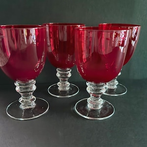May include: Four ruby red wine glasses with clear, decorative stems and bases. The glasses have a rounded bowl shape and are arranged on a dark surface. The glasses are made of glass.