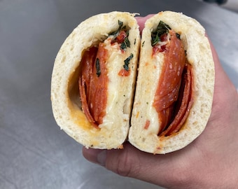 May include: A halved sandwich with pepperoni, melted cheese, and green herbs inside a light-coloured bread roll. The cut reveals the filling, highlighting the ingredients.