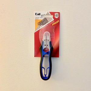Puede incluir: Embalaje rojo y blanco con un cortador de hilo azul y negro. El embalaje dice "Thread Clips" y "Kai Original".